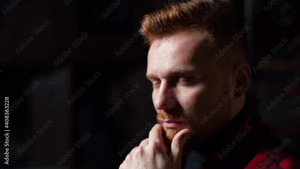 Close-up face of confident beared handsome young man thoughtfully holds hand near chin and looking away. Closeup of successful thinking male at indoor with dark interior. Shooting in slow motion. Close-up face of confident beared handsome young man thoughtfully holds hand near chin and looking away. Closeup of successful thinking male at indoor with dark interior. Shooting in slow motion.