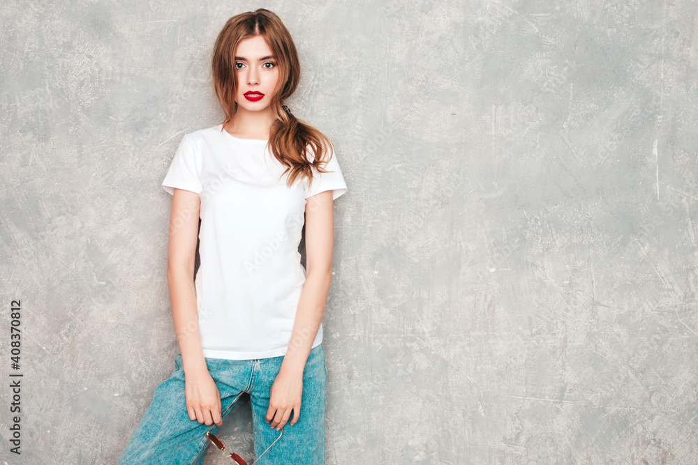 Young beautiful woman looking at camera. Trendy female in casual summer jeans clothes. Confident female with red lips.Model posing near gray wall in studio Young beautiful woman looking at camera. Trendy female in casual summer jeans clothes. Confident female with red lips.Model posing near gray wall in studio