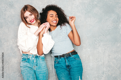 Two young beautiful smiling international hipster female in trendy summer jeans clothes. Sexy carefree women posing near gray wall in studio. Positive models having fun. Concept of friendship