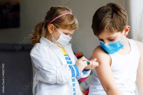 Little girl makes injection to brother, school kid boy. Children, siblings with medical mask playing doctor, holding syringe with vaccine. Coronavirus covid vaccination concept. Kids play role game