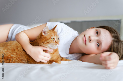 A girl with a ginger cat lies on a white bed. A young girl is stroking a ginger cat while lying on a white sheet