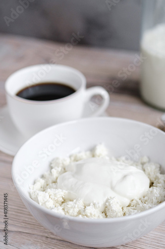 Cottage cheese with sour cream in a white bowl with coffee and milk on a white wooden table. Healthy breakfast. Rustic style.