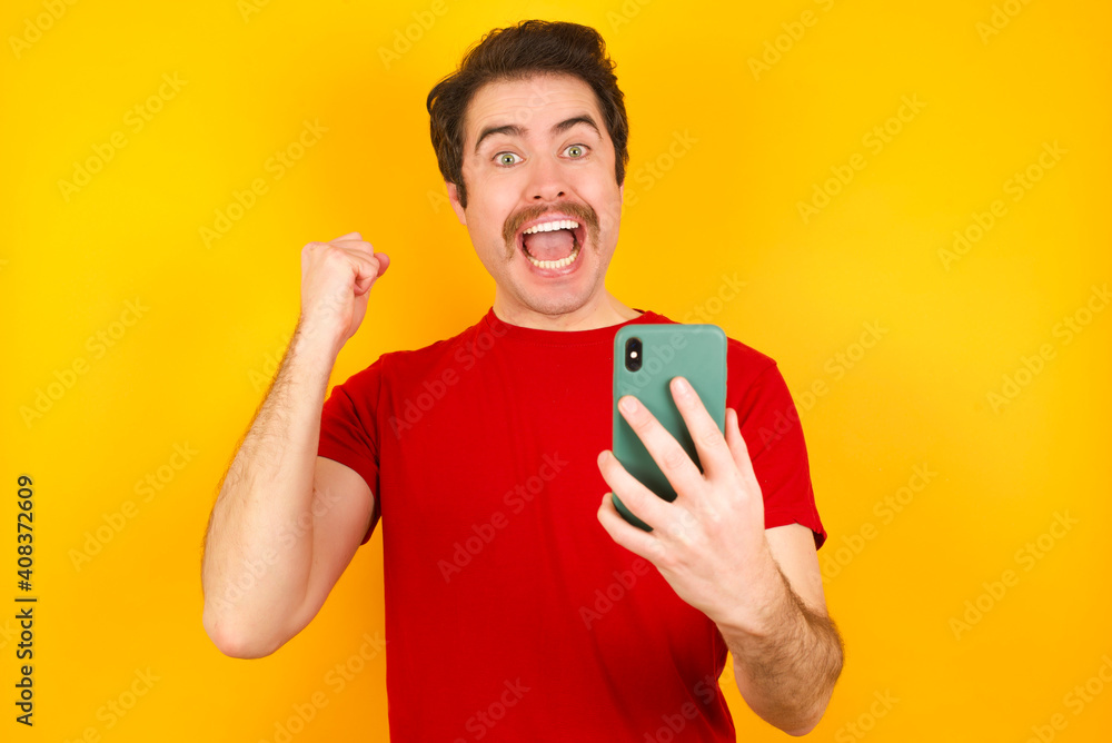 Portrait of Young Caucasian man wearing red t-shirt standing against yellow background holding in hands cell celebrating Portrait of Young Caucasian man wearing red t-shirt standing against yellow background holding in hands cell celebrating