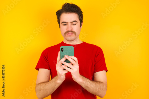Excited Young Caucasian man wearing red t-shirt standing against yellow background  winking and eye hold smart phone use read social network news © Roquillo