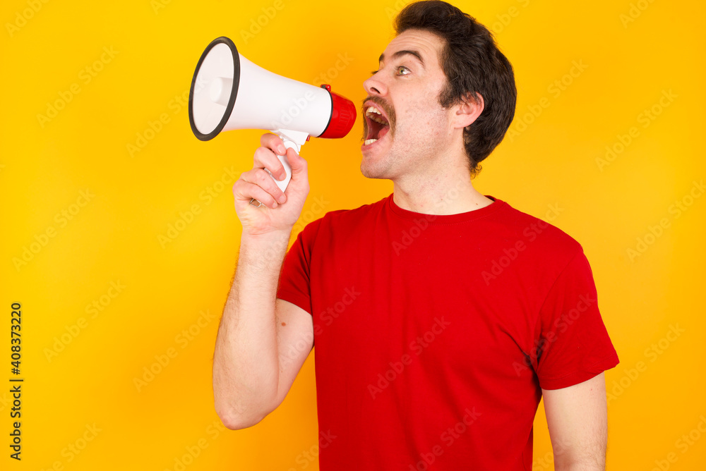 Young Caucasian man wearing red t-shirt standing against yellow background Through Megaphone with Available Copy Space Young Caucasian man wearing red t-shirt standing against yellow background Through Megaphone with Available Copy Space