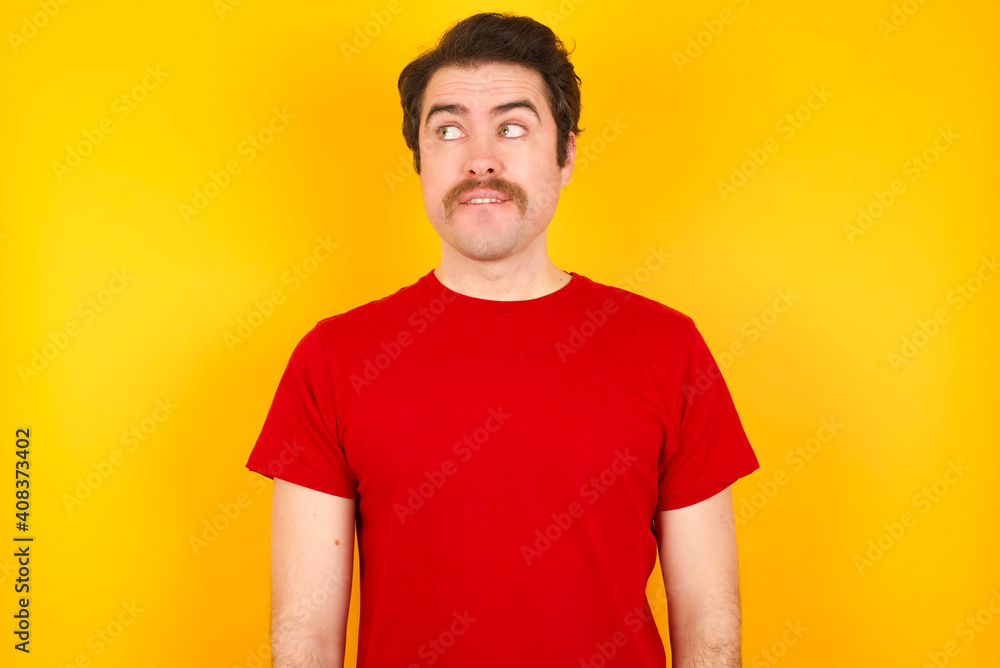 Obraz premium Amazed Young Caucasian man wearing red t-shirt standing against yellow background bitting lip and looking tricky to empty space.