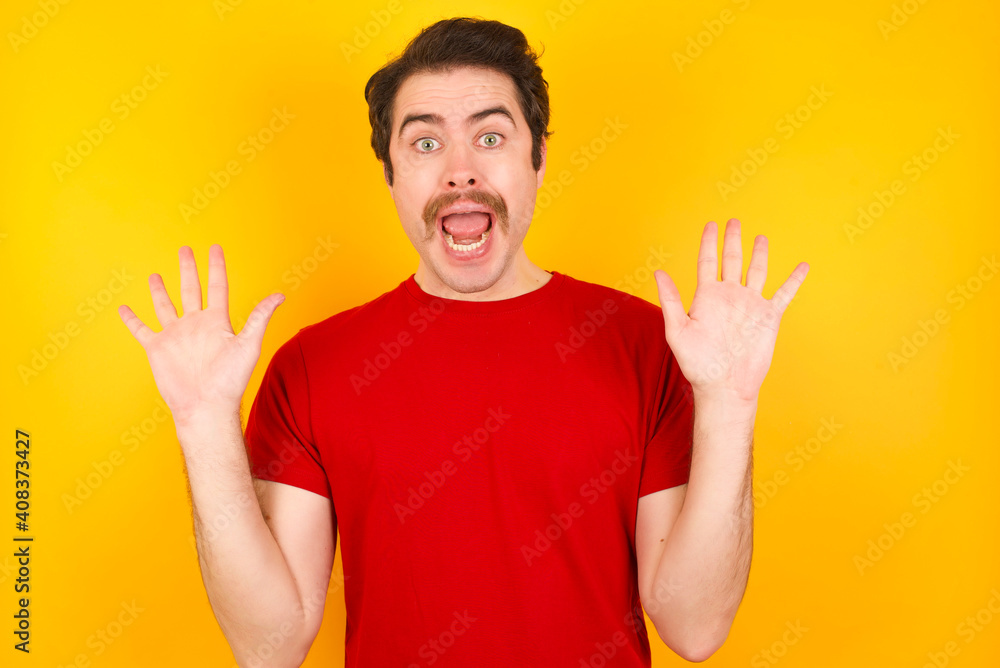 Optimistic Young Caucasian man wearing red t-shirt standing against yellow background raises palms from joy, happy to receive awesome present from someone, shouts loudly, Excited model screaming. Optimistic Young Caucasian man wearing red t-shirt standing against yellow background raises palms from joy, happy to receive awesome present from someone, shouts loudly, Excited model screaming.