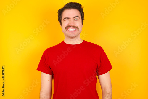 Coquettish Young Caucasian man wearing red t-shirt standing against yellow background smiling happily, blinking at camera in a playful manner, flirting with you.