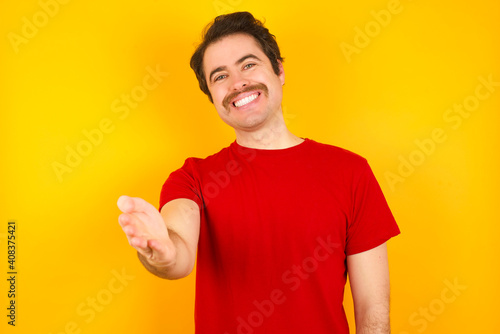Young Caucasian man wearing red t-shirt standing against yellow wall smiling friendly offering handshake as greeting and welcoming. Successful business.