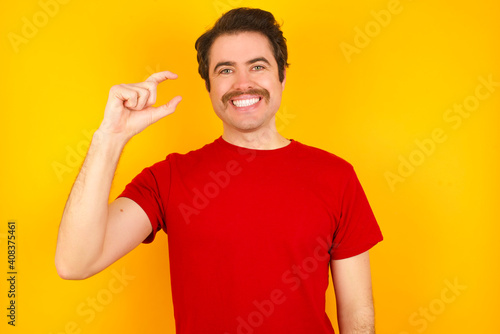 Young Caucasian man wearing red t-shirt standing against yellow wall smiling and confident gesturing with hand doing small size sign with fingers looking and the camera. Measure concept