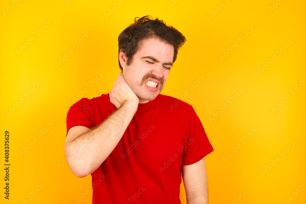 Young Caucasian man wearing red t-shirt standing against yellow wall Suffering of neck ache injury, touching neck with hand, muscular pain Young Caucasian man wearing red t-shirt standing against yellow wall Suffering of neck ache injury, touching neck with hand, muscular pain