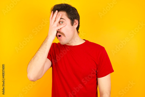 young Caucasian man wearing red t-shirt standing against yellow wall peeking in shock covering face and eyes with hand  looking through fingers with embarrassed expression.