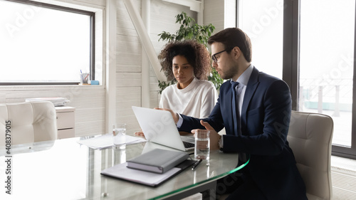Confident mentor businessman teaching African American businesswoman in office, pointing on laptop screen, helping with corporate software, diverse colleagues working on online project together