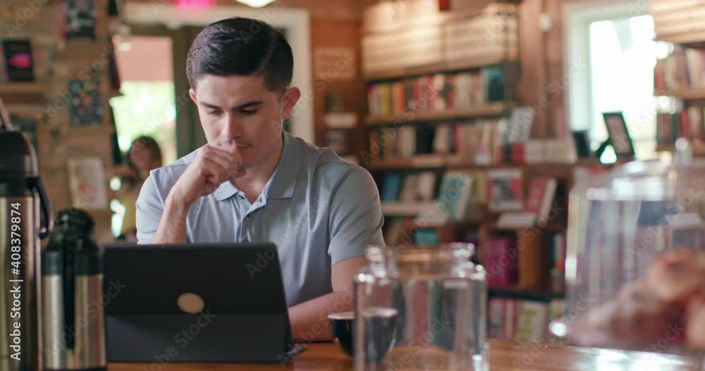 Portrait of young man sitting at table in coffee shop bookstore using a tablet, while a patron walks around the background. Portrait of young man sitting at table in coffee shop bookstore using a tablet, while a patron walks around the background.