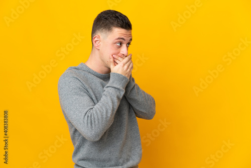 Young caucasian man isolated on yellow background covering mouth and looking to the side © luismolinero
