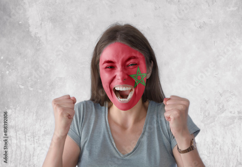 Young woman with painted flag of Morocco and open mouth looking energetic with fists up