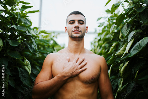 Handsome shirtless man standing with hand on chest amidst plants at greenhouse photo