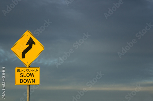 Blind corner slow down road sign against dark clouds