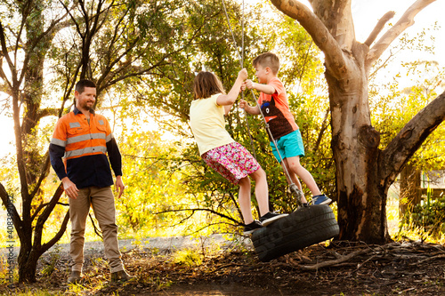 Dad playing with children in backyard on tyre swing in the afternoon