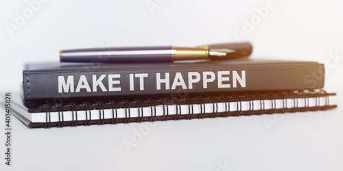 On the table are a notebook, a pen and a book. The book says - MAKE IT HAPPEN
