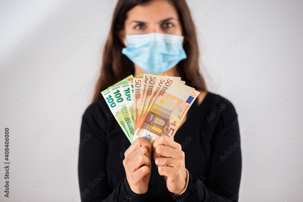 Happy woman holding multiple banknotes and wearing face mask in studio. Portrait of cheerful female adult with long brown hair with money in her hands. Happy woman holding multiple banknotes and wearing face mask in studio. Portrait of cheerful female adult with long brown hair with money in her hands.