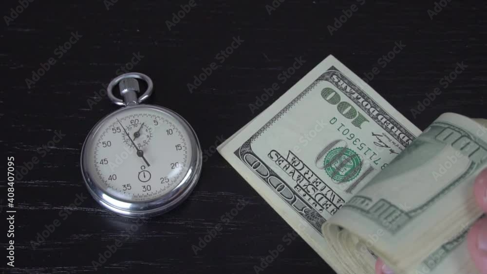 The stopwatch is on the table. Next to it, a hand drops a roll of 100 dollars bills, and the needle ticks. The concept of sales, discounts and investments in stocks and businesses. The stopwatch is on the table. Next to it, a hand drops a roll of 100 dollars bills, and the needle ticks. The concept of sales, discounts and investments in stocks and businesses.