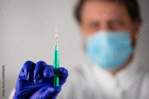 A doctor wearing a blue surgical face mask and blue gloves holding a disposable syringe with transparent injection solution - coronavirus vaccine