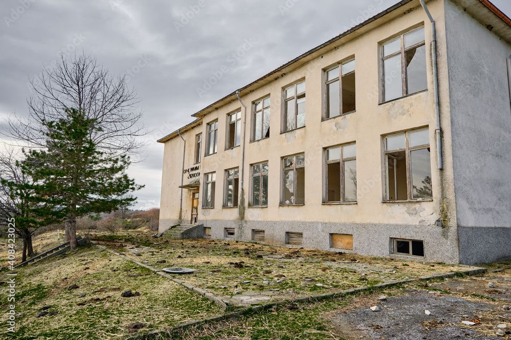 07.01.2021. Bulgaria, Kardzali. Old brownfield and abandoned soviet type school with overcast and very cloudy sky. School inside the green grass with broken windows.