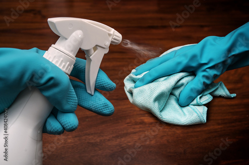 Hands in blue rubber gloves with a cleaning cloth and antibacterial sanitize spray bottle disinfecting a dark wooden surface to prevent covid-19 infection