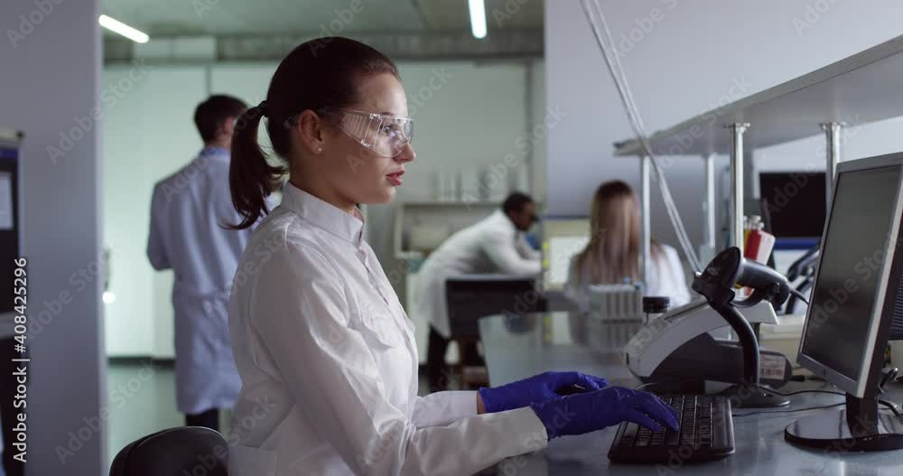 Portrait of busy concentrated Caucasian young pretty microbiology specialist in white coat and goggles in laboratory working browsing on computer examining test results doctors on background Side view Portrait of busy concentrated Caucasian young pretty microbiology specialist in white coat and goggles in laboratory working browsing on computer examining test results doctors on background Side view