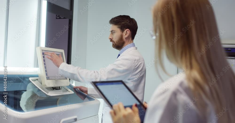 Over shoulder view. Rear of Caucasian young woman medical assistant standing in clinic lab room tapping on tablet while male professional doctor working typing on computer equipment during diagnosis Over shoulder view. Rear of Caucasian young woman medical assistant standing in clinic lab room tapping on tablet while male professional doctor working typing on computer equipment during diagnosis