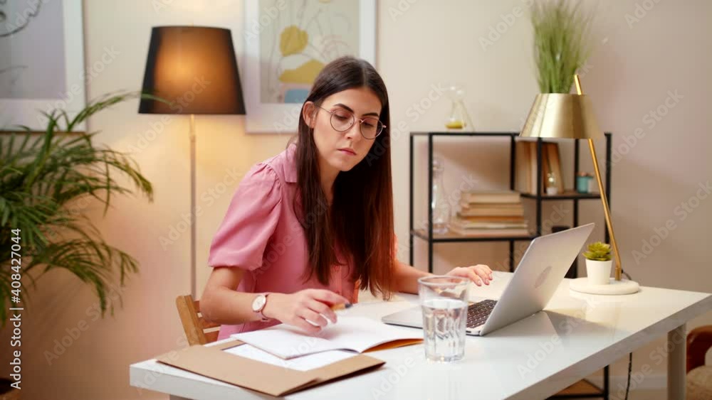 Smiling Young Business Woman Standing In Home Office At White Desk Working Alone On A Laptop, Browses The Internet, Writing Notes, Planning With Online Calendar, Bright Warm Light In Room Smiling Young Business Woman Standing In Home Office At White Desk Working Alone On A Laptop, Browses The Internet, Writing Notes, Planning With Online Calendar, Bright Warm Light In Room