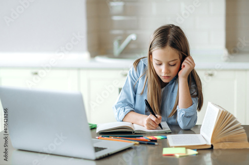 Distance learning, homeschooling during quarantine. Focused caucasian pretty schoolgirl doing her homework at home using laptop, sitting at the work desk
