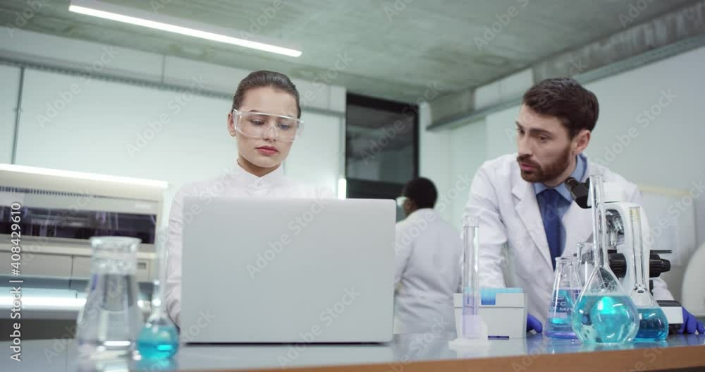 Portrait of Caucasian team of lab scientists male and female specialists working in laboratory together conducting experiment. Woman specialist typing on laptop computer while man using microscope Portrait of Caucasian team of lab scientists male and female specialists working in laboratory together conducting experiment. Woman specialist typing on laptop computer while man using microscope