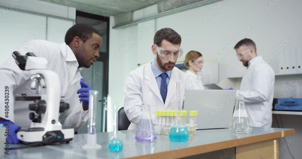 Portrait of busy African American male lab specialist in hospital laboratory looking in microscope doing medical research speaking with Caucasian colleague who is typing on laptop, developing cure Portrait of busy African American male lab specialist in hospital laboratory looking in microscope doing medical research speaking with Caucasian colleague who is typing on laptop, developing cure