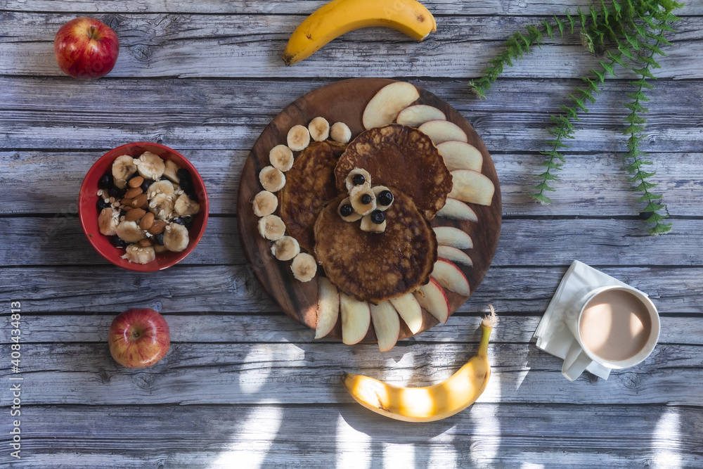 Fototapeta premium desayuno por la mañana un plato de madera con hot cakes con rebanadas de manzana y bananas alrededor con un tazon de frutas con una taza de cafe