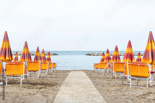 Fototapeta Naklejka Na Ścianę i Meble -  Lounge chairs on sand beach in Italy. Beach lounge area with orange umbrellas and green sunbeds. View on ocean and horizon. Sunbeds and parasols on the seashore. Adriatic coast, Rimini