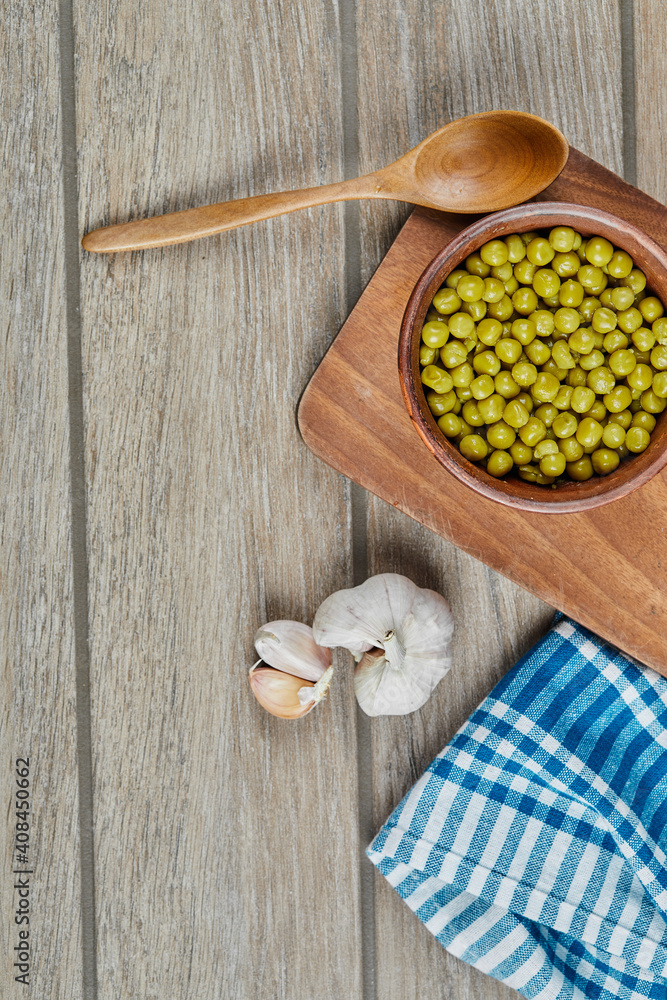 A bowl of boiled green peas with a spoon, garlic, and a blue tablecloth on a wooden table A bowl of boiled green peas with a spoon, garlic, and a blue tablecloth on a wooden table