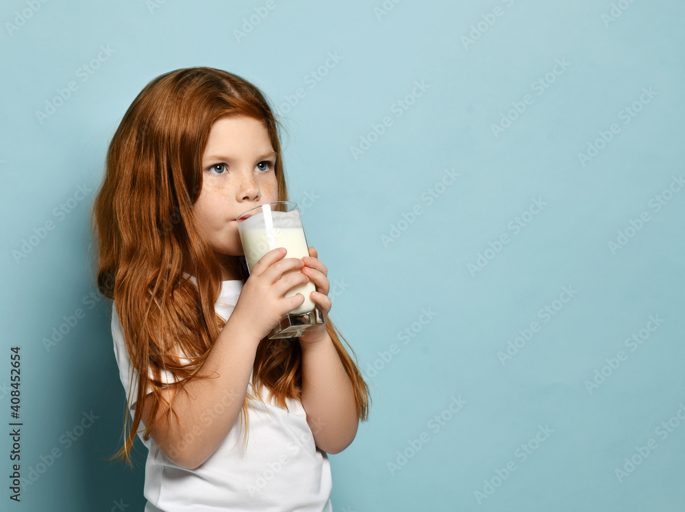 Portrait of red haired kid girl drinking milk or kefir yogurt looking at free copy space over light blue background. Health and diet concept. Healthy drink breakfast and clean food concept, copy space Portrait of red haired kid girl drinking milk or kefir yogurt looking at free copy space over light blue background. Health and diet concept. Healthy drink breakfast and clean food concept, copy space