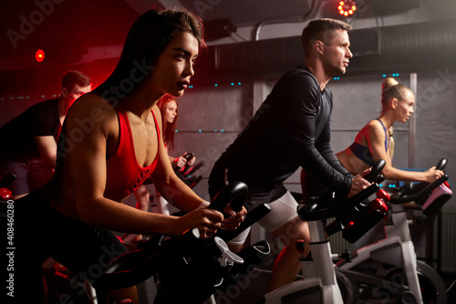 men and women biking in gym, exercising legs doing cardio workout cycling bikes, spinning in health club, wearing tracksuit sportive outfit