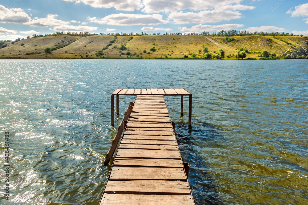 Wooden pier for fishing and swimming on bank of sunny river with hill slope on other side