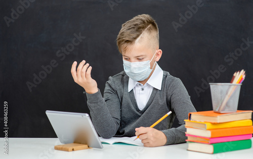 Teen boy wearing mask uses tablet computer at school near blackboard during corona virus and flu outbreak