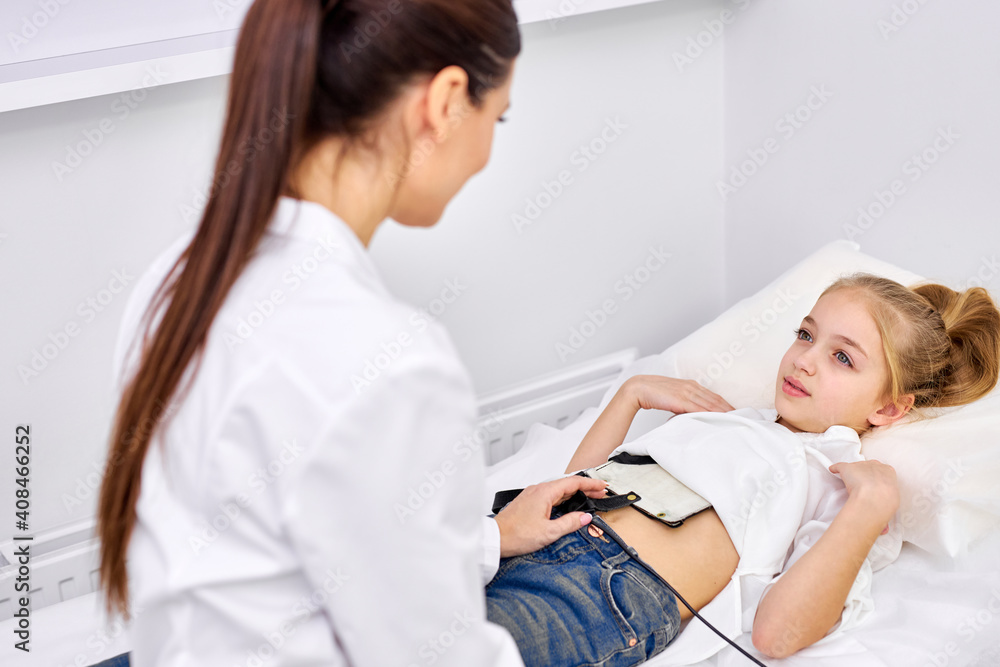 Fototapeta premium rear view on doctor putting special equipment on girl's belly for treatment, caucasian child lie on bed in hospital office, in a good mood. medicine and healthcare concept. focus on girl