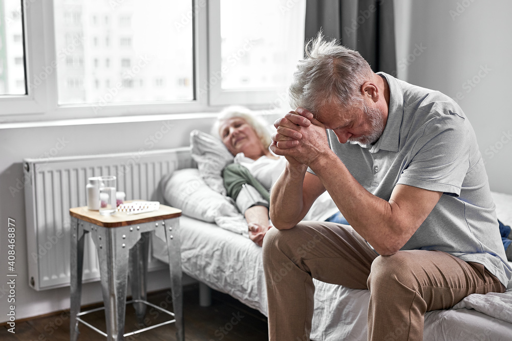 senior man sits with her sick ill wife lying on bed, feeling bad, female is at death's door, man is very worried about her senior man sits with her sick ill wife lying on bed, feeling bad, female is at death's door, man is very worried about her