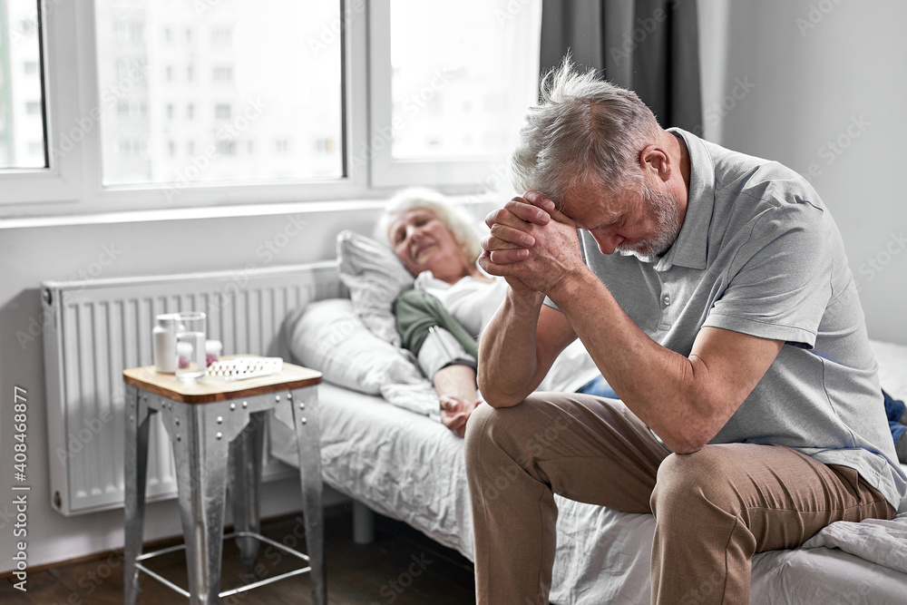 senior man sits with her sick ill wife lying on bed, feeling bad, female is at death's door, man is very worried about her