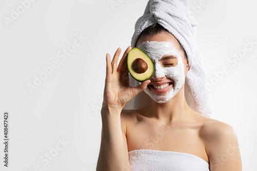 Overjoyed young pretty female with mask on face in white towel cover eyes by avocado.