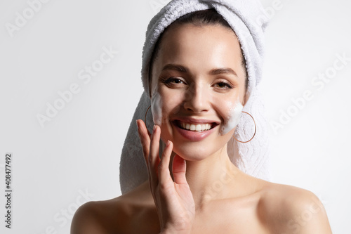 Young joyful girl applying face wash foam on cheek, feeling excited of skin condition.