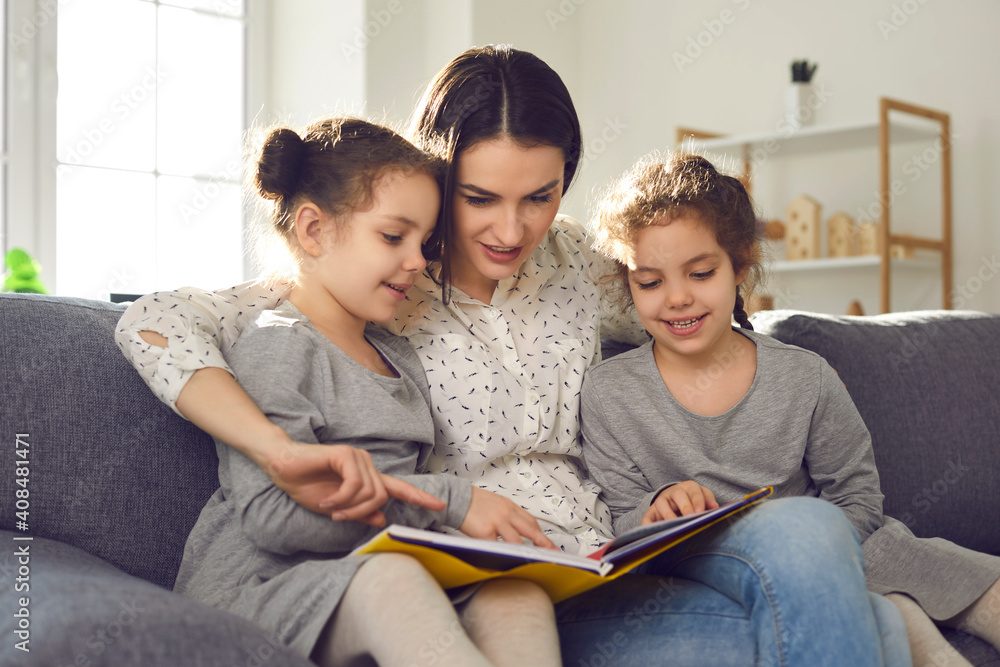 Happy young mother reading a book sitting at home on the couch with her twin daughters. Woman hugs children and tells children funny tales and stories. Concept of child development. Happy young mother reading a book sitting at home on the couch with her twin daughters. Woman hugs children and tells children funny tales and stories. Concept of child development.