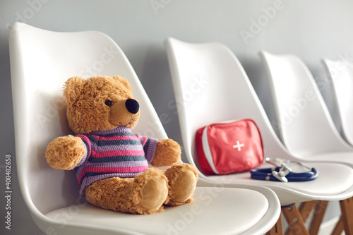 First-aid kit, stethoscope and cute teddy bear on white chairs in waiting room of children's medical center or pediatric clinic. Concept of kids' visit to hospital to see pediatrician or family doctor photo