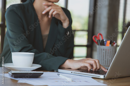 Businesswoman working on laptop in her workstation.Black blank screen smartphone, potted plant, pencil, notes, earphone on wooden desk. Close up
